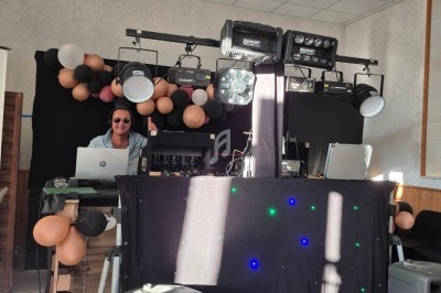 Un DJ mixe derrière une table équipée de matériel audio et d'éclairage, décorée de ballons dans une salle.