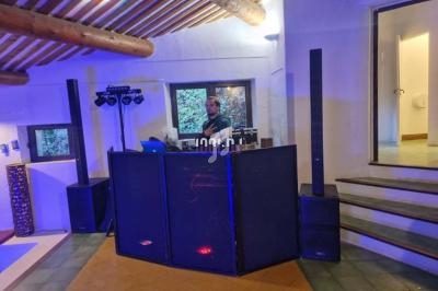 Un DJ mixe derrière une table équipée de matériel audio et éclairée par des lumières colorées dans une pièce avec poutres…
