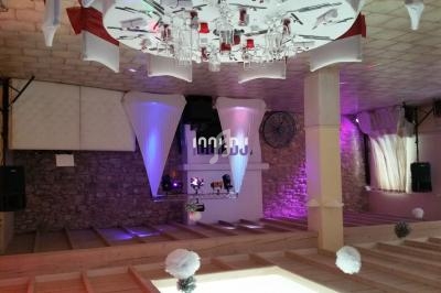 Salle de réception décorée avec des tables dressées, éclairages colorés et espace DJ en arrière-plan.