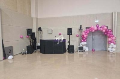 Salle décorée avec des ballons roses et blancs, équipée de matériel audio et d'une arche près d'une porte grise.