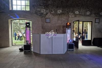 Poste de DJ avec enceintes et éclairages dans une salle en pierre, portes ouvertes sur un jardin avec des invités.