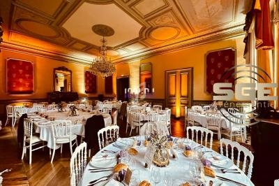 Salle de réception élégante avec tables dressées, chaises blanches et éclairage chaleureux sous un plafond orné.