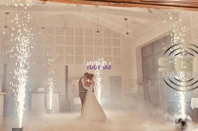 Un couple de mariés danse dans une salle éclairée par des étincelles et enveloppée de fumée.