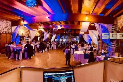 Salle de réception décorée, éclairage coloré, invités assis à des tables rondes, vue depuis une régie musicale.