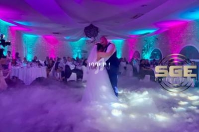 Un couple danse leur première danse de mariage dans une salle illuminée de lumières colorées, entourée de fumée au sol.