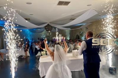 Une mariée lève les bras devant des invités dans une salle décorée, entourée d'effets pyrotechniques.