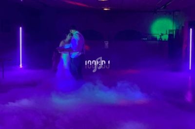 Un couple danse dans une salle sombre éclairée par des lumières colorées et entourée de fumée au sol.