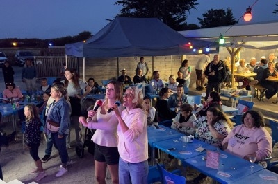 Des personnes participent à une soirée karaoké en plein air, entourées d'un public assis sous un éclairage coloré.