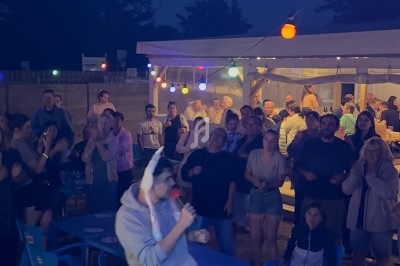 Un homme chante au micro devant un public assis et debout, éclairé par des guirlandes lumineuses en soirée.