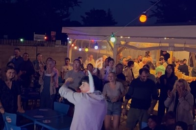 Un homme avec un micro anime une soirée en plein air devant un public assis et debout sous des guirlandes lumineuses.