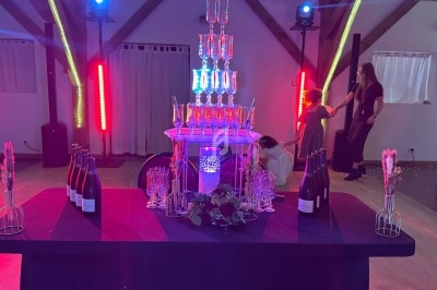 Tour de flûtes à champagne illuminée sur une table décorée, entourée de bouteilles et de lumières colorées.