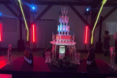Tour de coupes en verre illuminée, entourée de bouteilles et décorations, dans une salle avec éclairage coloré.