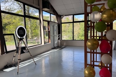 Salle lumineuse avec de grandes baies vitrées, un trépied avec un anneau lumineux et une structure décorée de ballons.