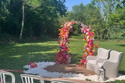 Arche florale colorée avec fauteuils en osier, installée en extérieur sur une pelouse entourée d'arbres.
