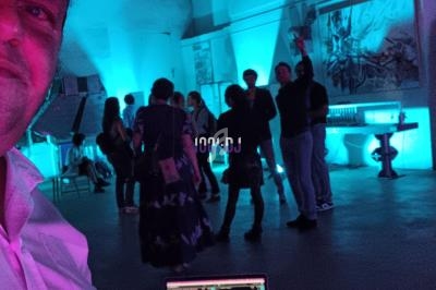 Un DJ mixe sur son ordinateur dans une salle éclairée de lumières bleues, avec des personnes discutant en arrière-plan.