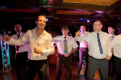 Des hommes en chemise dansent ensemble dans une salle éclairée, avec un plafond en bois sombre.