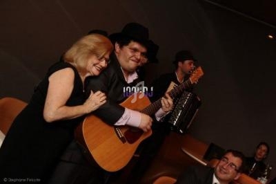 Un homme joue de la guitare entouré d'une femme souriante et d'autres musiciens en arrière-plan.