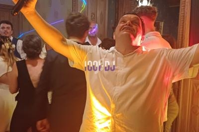 Un homme en chemise blanche chante avec enthousiasme dans une salle festive, entouré de personnes dansant.