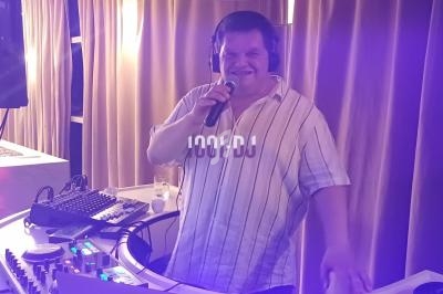 Un DJ souriant mixe de la musique avec un micro à la main, devant une table de mixage éclairée dans une ambiance tamisée.