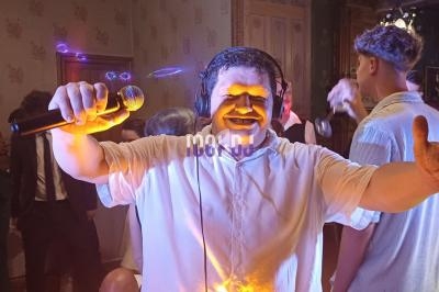 Homme souriant avec un micro et un casque audio, levant le pouce, dans une ambiance festive avec des personnes en arrière…