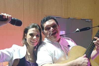 Une femme souriante tenant un micro et un homme jouant de la guitare, debout dans une salle éclairée.