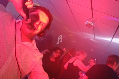 Homme chantant au micro dans une ambiance festive avec des lumières colorées et des personnes dansant en arrière-plan.