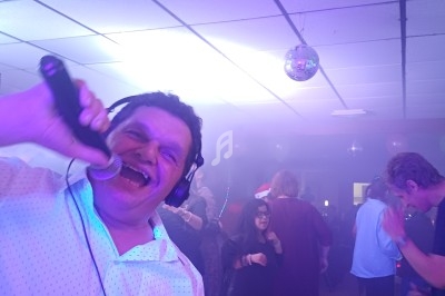 Un homme chante au micro avec un casque audio lors d'une fête animée dans une salle éclairée par des lumières colorées.