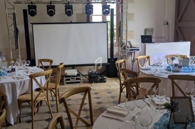 Salle de réception décorée avec des tables dressées, un écran de projection et un éclairage suspendu.