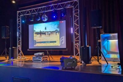 Scène équipée d'un écran projetant une vidéo, entourée de projecteurs, enceintes et matériel audiovisuel.