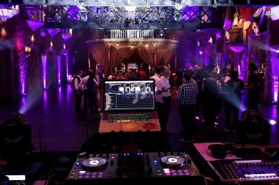 Vue depuis une table de mixage avec un ordinateur, dans une salle de réception éclairée, des invités dansent au fond.