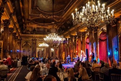 Salle de réception luxueuse avec chandeliers, colonnes dorées et invités dînant devant une scène avec musiciens.
