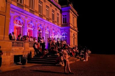 Façade d'un bâtiment éclairé en violet avec des personnes assises et debout sur les marches, de nuit.