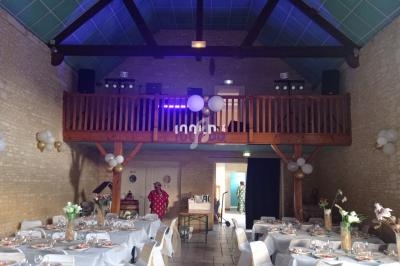 Salle de réception décorée avec des tables dressées, des ballons et une mezzanine en bois équipée de matériel audio.