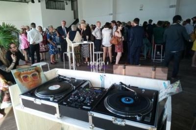 Deux platines vinyles et une table de mixage au premier plan, avec des invités debout dans une salle en arrière-plan.