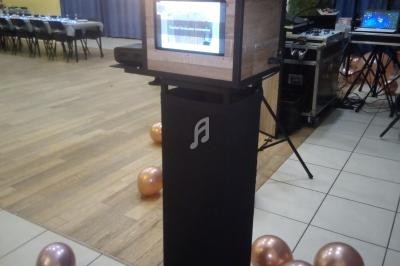 Borne photo en bois et métal dans une salle décorée de ballons, avec tables dressées en arrière-plan.
