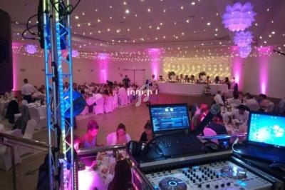 Salle de réception décorée avec des lumières roses, tables dressées et équipement de DJ au premier plan.
