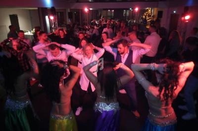 Des personnes dansent en groupe avec des danseuses en tenue colorée dans une salle éclairée par des lumières tamisées.