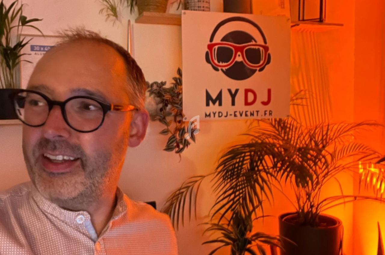 Homme souriant portant des lunettes devant un mur décoré avec des plantes et une affiche ’MYDJ’.