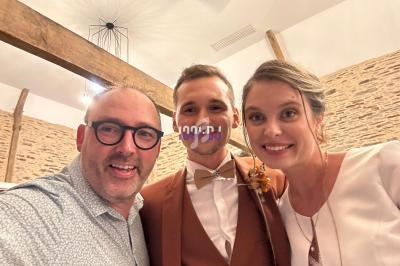 Un homme prend un selfie avec un couple souriant dans une salle aux murs en pierre et poutres apparentes.