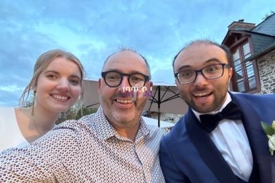Un homme prend un selfie avec une femme en robe blanche et un homme en costume bleu, souriants devant un ciel nuageux.