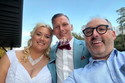 Un couple en tenue de mariage pose avec un homme souriant pour une photo en extérieur, sous un ciel dégagé.