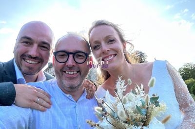 Un homme et une femme en tenue de mariage sourient avec un ami, bouquet de fleurs séchées à l'avant-plan.