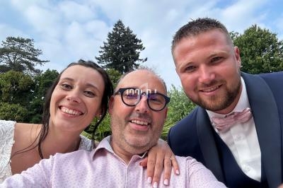 Un homme souriant prend un selfie avec une femme en robe blanche et un homme en costume devant des arbres.