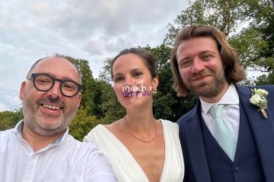 Un homme prend un selfie avec une mariée en robe blanche et un marié en costume bleu, dans un cadre extérieur verdoyant.