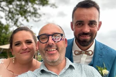 Un homme souriant prend un selfie avec une femme en robe claire et un homme en costume bleu devant un paysage extérieur.