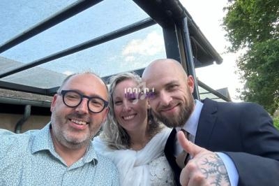 Trois personnes souriantes posent ensemble à l'extérieur sous une structure vitrée, l'une fait un signe de pouce levé.