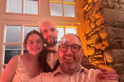 Un couple en tenue de mariage pose avec un homme souriant devant un mur en pierre et une fenêtre éclairée.