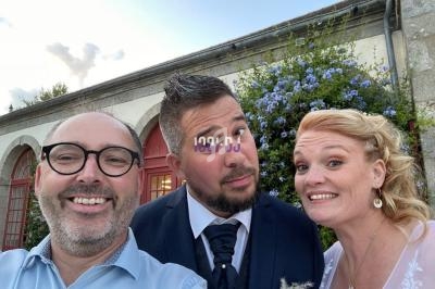 Trois personnes souriantes posent devant un bâtiment en pierre avec des fleurs bleues en arrière-plan.