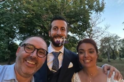 Un homme en costume et une femme en robe blanche posent avec un autre homme souriant devant des arbres au crépuscule.