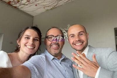Un couple souriant pose avec un homme au centre, l'époux montre son alliance dans un intérieur décoré.